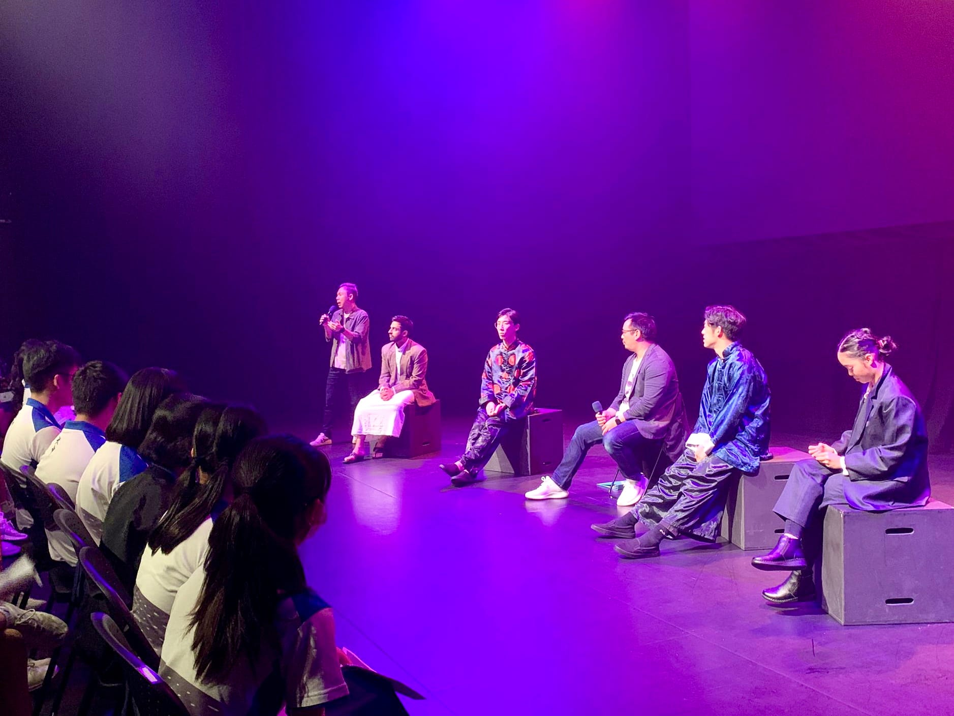 Six people on a purple-lit stage, one standing with a mic, five seated on boxes, facing a seated audience.